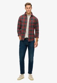 Rood en groen geruit flanellen shirt over een lichtgrijs T-shirt, gecombineerd met blauwe spijkerbroeken en beige sneakers. Gepresenteerd in een staande pose.