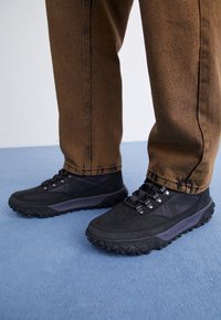 Chaussures noires d'extérieur avec un tissu texturé, des semelles en caoutchouc et des accents gris, associées à un pantalon marron, posées sur une surface bleue.