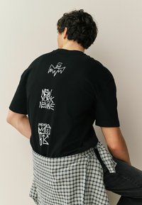 Göndör sötét hajú férfi fekete grafikus pólóban és csípője köré kötött kockás ingben, egyszínű háttér előtt ülve.