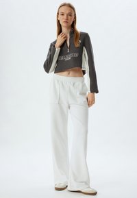 Cazadora gris recortada con estampado de "UNLIMITED" y acentos en blanco, combinada con pantalones anchos blancos que tienen bolsillos frontales y una cintura elástica.