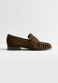 Mocassino in suede marrone con tacco basso impilato, cucitura a mocassino e una strap decorativa sulla tomaia, mostrato di profilo su uno sfondo bianco.