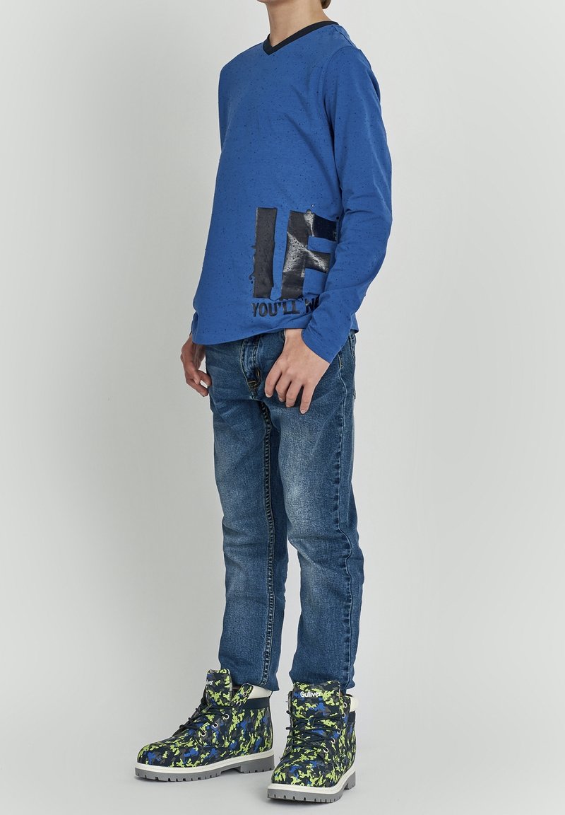 Barn iført blå langærmet skjorte, blå jeans og mønstrede grønne og blå vandrestøvler, stående mod en ensfarvet baggrund.