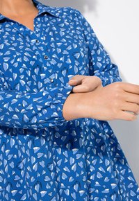 Blå knapp-up skjorta med ett hjärt-mönster i ljusblått, med rynkade ärmar och en krage. Bomullstyg med en mjuk textur.