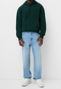 Felpa verde scuro con tasca frontale, abbinata a jeans dritti blu chiaro e sneaker bianche. Design semplice e casual.