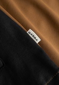 Gros plan d’un tissu noir et marron avec une étiquette de marque blanche GABBA visible cousue entre les deux textiles.