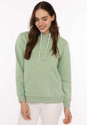 Junge Frau lächelt, trägt einen hellgrünen Hoodie mit Fronttasche und weißem Text-Kreis-Design, kombiniert mit weißen Hosen, vor weißem Hintergrund.