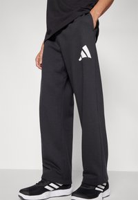 Mørkegrå sweatpants med løs pasform, der har et hvidt logo på venstre lår. Stoffet ser blødt ud og er kombineret med sorte sneakers.
