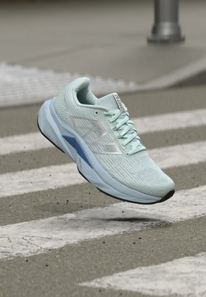 Gaismi zilas krāsas sporta apavi ar "N" logotipu, pacelti virs pārejas uz asfalta ceļa.