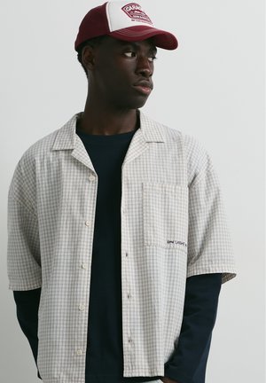 Homme portant une casquette bordeaux et blanche, une chemise à manches courtes à carreaux beige clair sur un t-shirt noir à manches longues, regardant vers sa gauche sur un fond uni.