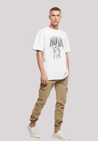 Hvit grafisk t-skjorte med "HULK" trykk og tegneserie-neve, sammen med beige cargo-bukser med flere lommer, og hvite sneakers.