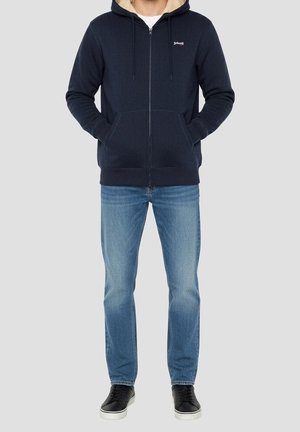Mand iført marineblå lynlåstrøje med hætte og beige for, blå jeans og sorte sneakers, stående med hænderne i jakke-lommerne.