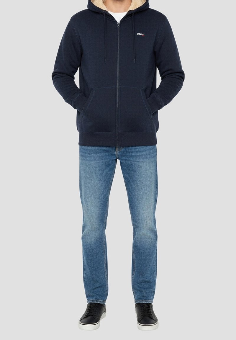 Homme portant une veste à capuche zippée bleu marine avec doublure beige, jean bleu et baskets noires, debout les mains dans les poches de la veste.