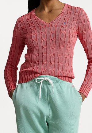 Femme portant un pull rose à col en V en maille torsadée et un pantalon de survêtement vert menthe, les mains dans les poches, sur fond uni.