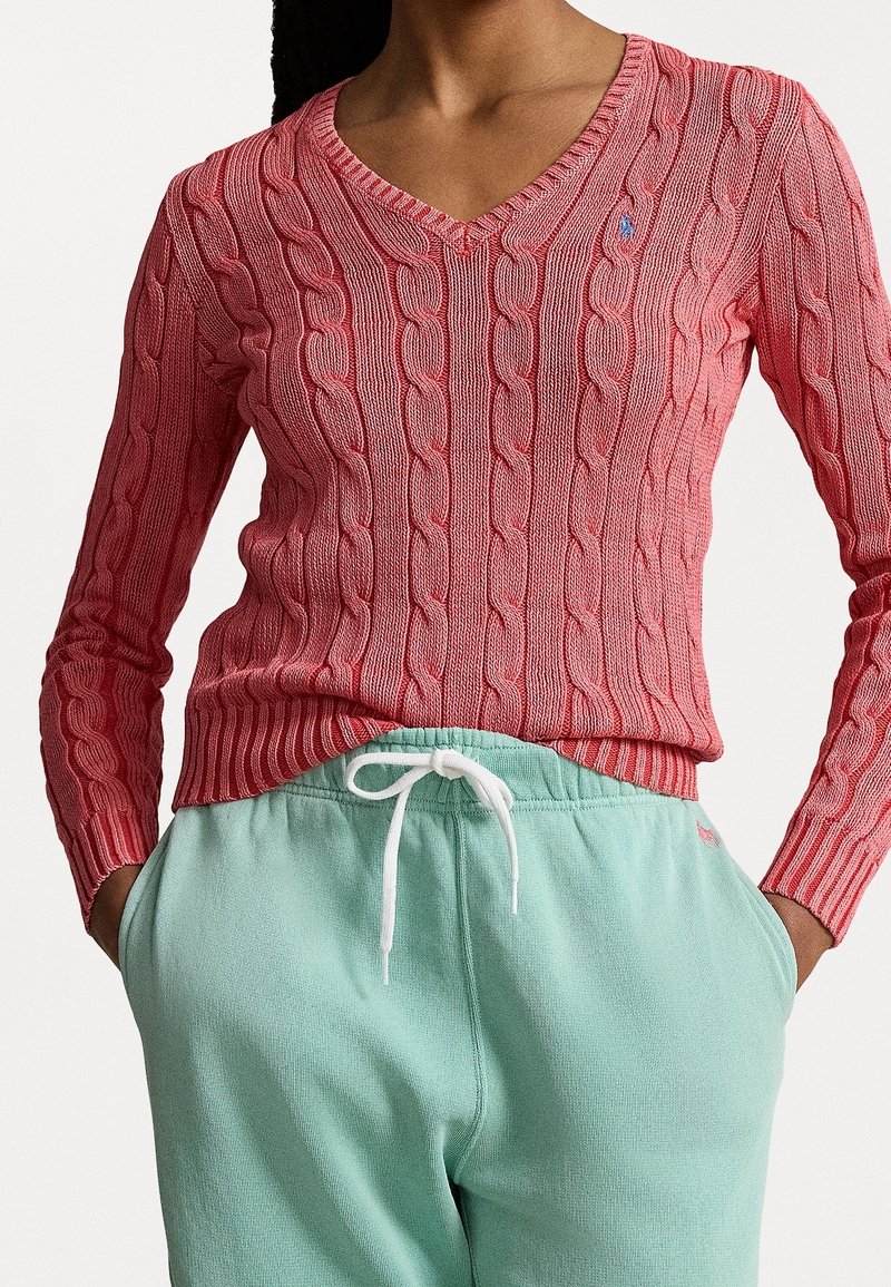 Femme portant un pull rose à col en V en maille torsadée et un pantalon de survêtement vert menthe, les mains dans les poches, sur fond uni.