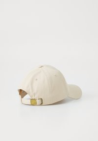 Beige bomullskeps med en böjd skärm, justerbart band med en guldfärgad metallspänne på baksidan och sex ventilationshål.