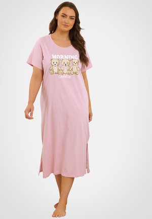 Kvinde iført en lyserød lang natkjole med tre bamser og teksten "Morning Cuddles," stående barfodet mod en ensfarvet baggrund.