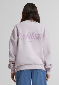 Rosa oversized hettegenser med "Drohited" i lilla kursiv tekst på baksiden. Har ribbestrikkede mansjetter og liv, myk stofftekstur.