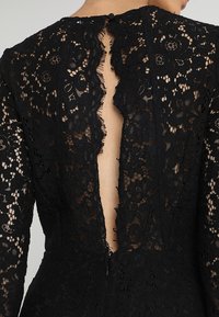 Robe en dentelle noire avec des motifs floraux complexes, manches longues, dos en V profond et fermeture à boutons au niveau du cou.