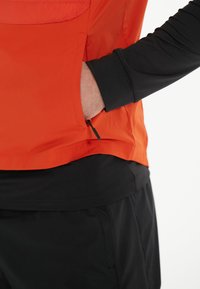 Orange lättviktig jacka med en dragkedjeficka, ihop med ett svart långärmat baslager. Slät textur, modern design.