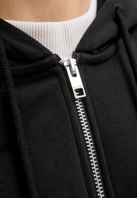 Schwarze kapuzenjacke mit Reißverschluss, ausgestattet mit einem silbernen Reißverschluss, strukturiertem Stoff und einem weißen gerippten Kragen. Akzente beinhalten einen geprägten Reißverschlussanhänger.