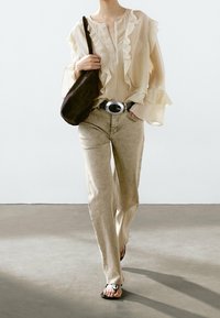Beige frilled blouse with long sleeves, light faded jeans, and black belt with a large silver buckle; paired with black flip-flops and brown shoulder bag.