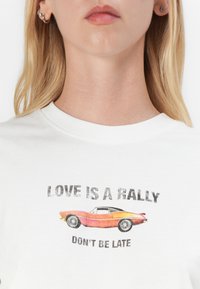 White cotton t-shirt featuring a graphic print of an orange car with text: "LOVE IS A RALLY DON'T BE LATE." Text has a distressed effect.
