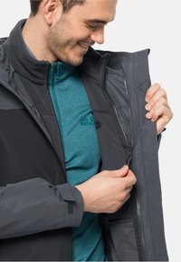 Heren grijze buitenjas met een gegroefde fleece voering, ritssluiting aan de voorkant en verstelbare manchetten. Draagt over een tealkleurig fleece shirt met logo detail.