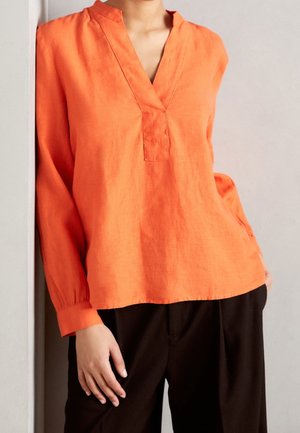 Persoon draagt een feloranje blouse met lange mouwen en V-hals en zwarte broek, staand tegen een lichtgekleurde muur.