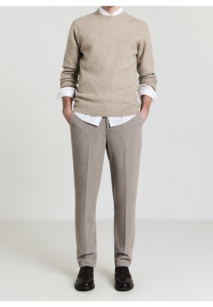 Mann trägt beigen Pullover über weißem Hemd, beige Hose und schwarze Schuhe, steht mit Händen in den Taschen vor hellem Hintergrund.