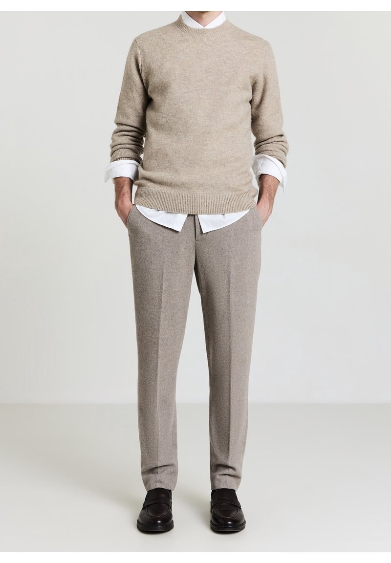 Homme portant un pull beige par-dessus une chemise blanche, un pantalon beige et des chaussures noires, debout les mains dans les poches devant un fond clair.