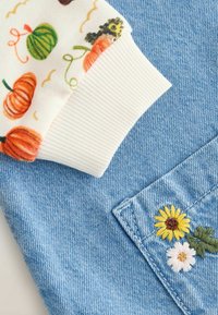 Tissu en denim bleu clair avec une poche décorative ornée de fleurs brodées jaunes et blanches. Poignets en crème avec des motifs de citrouilles colorées.