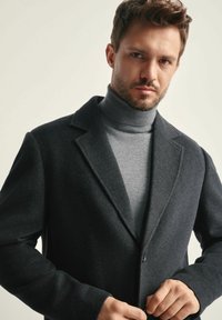 Mężczyzna zapinający ciemnoszary płaszcz na jasnoszary sweter z golfem, na neutralnym tle, patrzący bezpośrednio w kamerę.