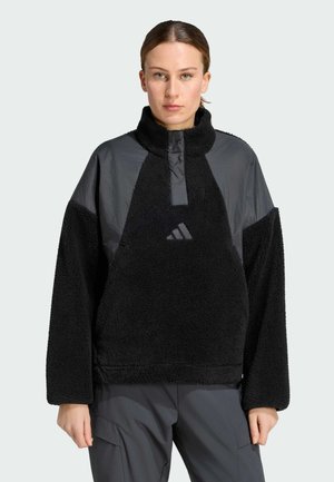 Mujer de frente, con un forro polar negro y gris de cuello alto y mangas largas, de pie frente a un fondo liso.