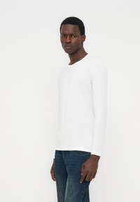 Μαύρος άνδρας όρθιος, φοράει απλό λευκό μακρυμάνικο πουκάμισο και μπλε τζιν, κοιτάζοντας ελαφρώς πλάγια μπροστά από ένα λευκό φόντο.