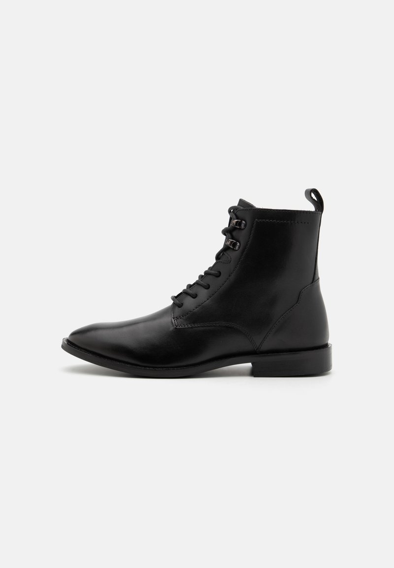 Bottine noire en cuir avec lacets, bout rond, petit talon et languette à l'arrière sur fond gris clair.