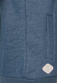 Felpa blu con tessuto testurizzato, tasca laterale e una toppa a forma di diamante bianco con un emblema vicino all'orlo.