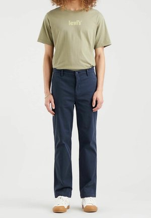 Persona che indossa una t-shirt Levi's verde oliva, pantaloni blu navy, sneakers bianche con suole di gomma, in piedi davanti a uno sfondo semplice.