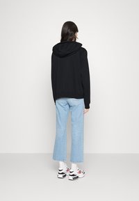 Sudadera negra con capucha, de corte relajado. Combinada con vaqueros de pierna ancha en azul claro y zapatillas blancas con detalles en rojo. Fondo neutro.