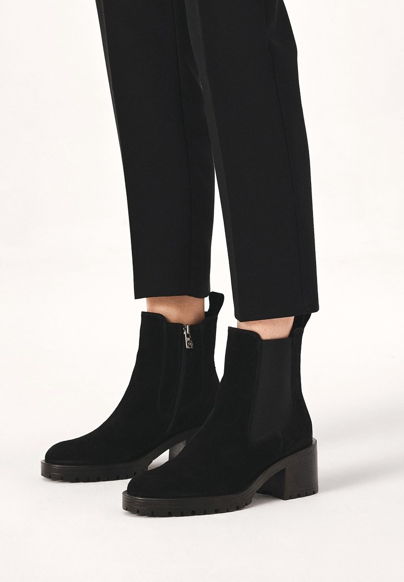 Bottines en daim noir avec talons carrés et fermetures éclair sur les côtés, portées avec un pantalon court noir sur un fond clair.
