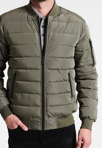 Chaqueta bomber acolchada verde oliva con cierre de cremallera en el frente, cuello acanalado, dos bolsillos laterales y un bolsillo con cremallera en la manga izquierda.