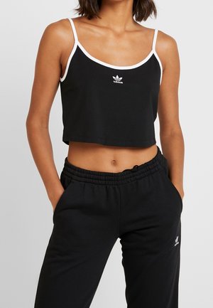Kobieta ubrana w czarny crop top Adidas z białym wykończeniem i pasujące czarne spodnie dresowe Adidas, ręce w kieszeniach, widziana od szyi do bioder.