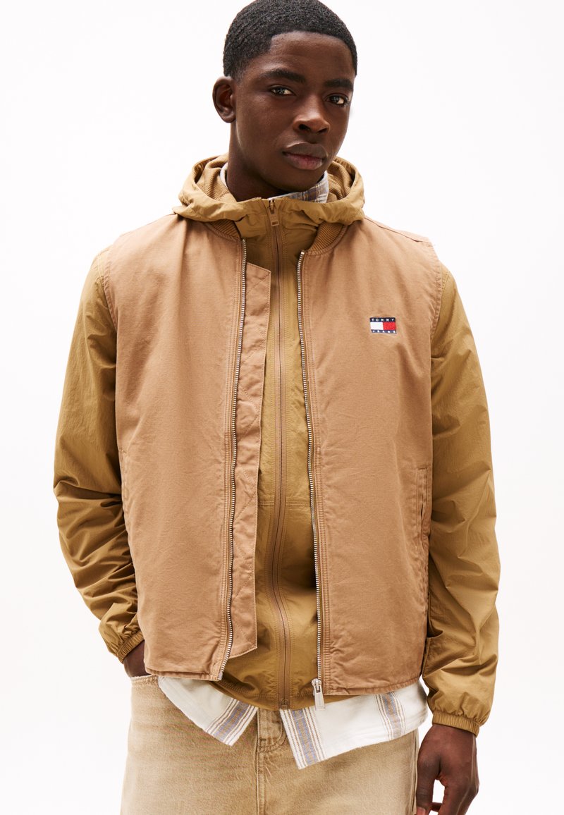 Lichtbruine, ritsjack die over een tan hoodie is gelaagd. Bevat een logopatch, elastische manchetten en een gladde textuur. Draag met een overhemd met kraag.