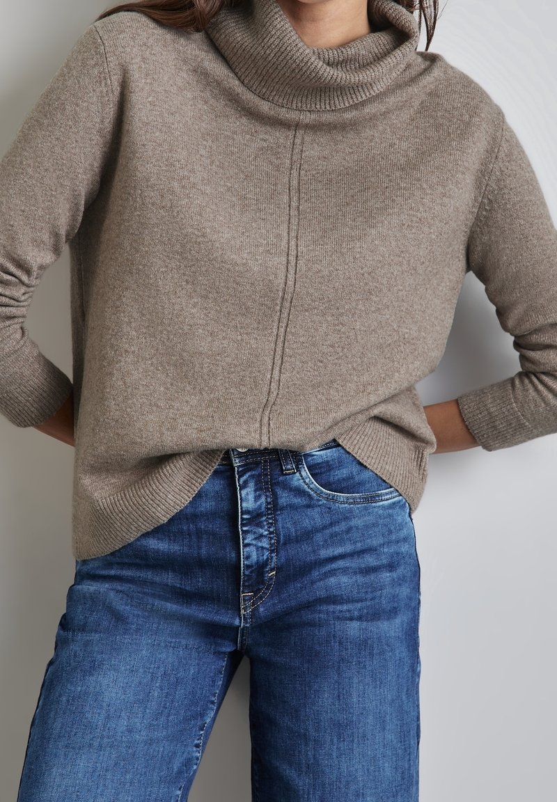 Pull à col roulé marron clair avec tricot texturé et col côtelé, associé à un jean en denim bleu à coupe classique avec un léger effet délavé.