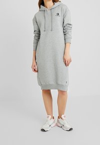Vestido sudadera gris con un bolsillo delantero, mangas largas y capucha con cordón. Presenta un pequeño logo negro en el pecho. Material suave.