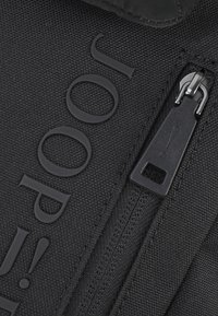 Czarny plecak z tkaniny o teksturowanej powierzchni, z wyraźnym logo, zapięciem na zamek oraz eleganckimi czarnymi detalami metalowymi.