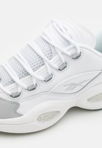 White athletic shoe featuring a mesh upper, leather accents, reflective details, prominent lacing system, and hexagonal textured sole.