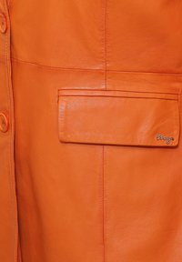 Oranje leren jas met een voorkantzak, ronde knopen en subtiele stiksels. De hardware omvat een merketiket op de zak.