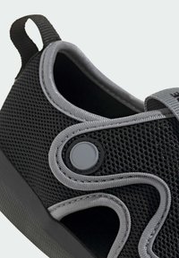 Svart mesh-sko med grå detaljer, en rund knappdetalj och en texturerad sula. Designen inkluderar utskurna sektioner för andningsförmåga.