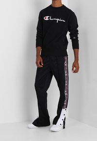 Felpa nera con logo "Champion" bianco, abbinata a pantaloni neri con strisce laterali e dettagli a snap. Sneakers bianche completano l'outfit.