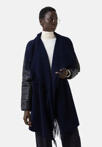 Námořnická modrá šála s třásněmi, vyrobená z měkkého materiálu; vrstvená přes světlý top s vysokým límcem, doplněná šedým rukávem.